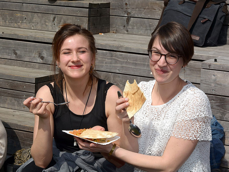 Zwei Frauen sind beim Essen der verschiedenen internationalen Fastesnspeisen zu sehen. Sie halten einen Teller mit einer Speise und ein Fladenbrot in der Hand